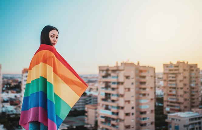 An Asian woman wrapped in a rainbow flag overlooks a city. Photo: Dark Saiyaman/Adobe Stock